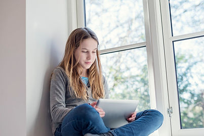 A teen girl sitting in her room smiling by her window. She is on her tablet talking with her therapist online. She is comfortable and happy in her own environment at home. Marble Wellness offers virtual child therapy for clients living in MO or IL!