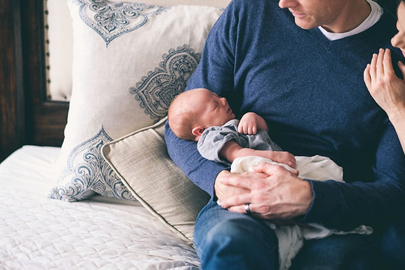 A dad holds his newborn baby while sitting on a bed. Therapy for men can help with the transition into parenthood. Marble Wellness in St. Louis, MO offers in-person therapy for men, online therapy in Missouri and Illinois, and park therapy.