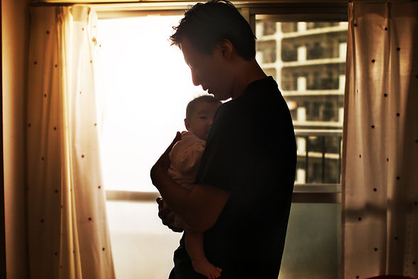 A man stands in front of a window holding his baby. Men can develop paternal postpartum depression and therapy for men in St. Louis can contribute to improvement. Marble Wellness specializes in therapy for dads in West County, MO.
