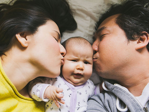 A mom and dad kiss their baby in bed. Couples therapy in St. Louis is offered at Marble Wellness and can help couples adjust to life with a new baby. Therapy for postpartum depression and anxiety in St. Louis is available at Marble Wellness.