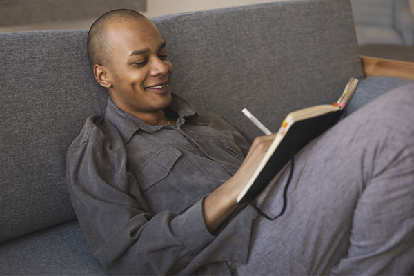Photo of a smiling man with chronic illnes sitting on the couch writing notes in a notebook after counseling for chronic illness in St. Louis, MO, or via online therapy in Missouri and online therapy in Illinois. You can get options for a chronic illness counselor near you, for therapy for chronic pain, anxiety, and more.