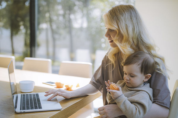 Photo of a mom working on the computer with her baby on her lap eating an orange. Therapy for moms in Chicago, Il | Therapy for working moms in Chicago, IL | overwhelmed mom | therapy for moms near me | online therapy for moms | 63122 | forrest glen 60630 | Lincoln Park is 60614 | north center 60613 | 60618.