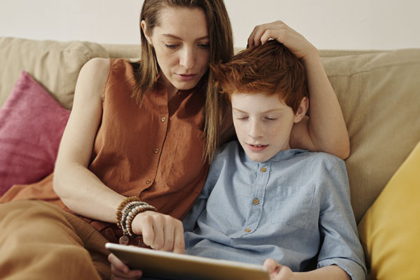 Photo of a mother and son sitting on the couch where the mom is helping her son learn a subject for school on a tablet. Online therapy for moms in St. Louis, MO | therapy for moms | overwhelmed moms.