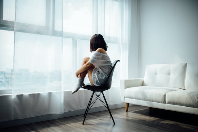 Woman curled up on a chair looking out a large window with sheer curtains representing grief and sadness. Marble Wellness is a therapy practice in Ballwin Mo offering grief counseling. Get started today.
