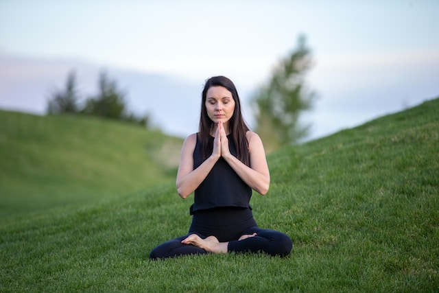 A woman in yoga clothes practices meditation on a grassy knoll. Self care is an important practice when handling grief. Schedule an appointment with a STL therapist today.