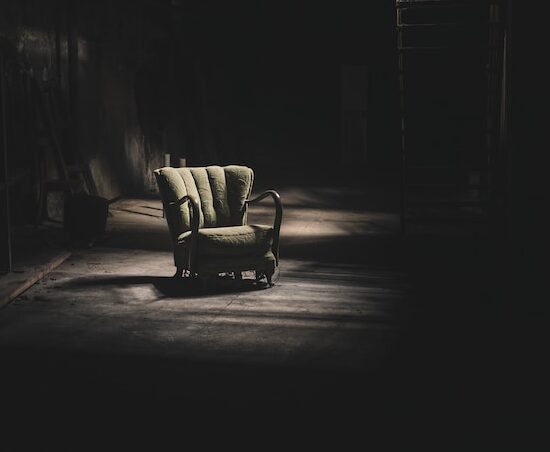 An old green chair sitting in the shadows of a dark room representing the feelings of isolation when dealing with grief & loss.