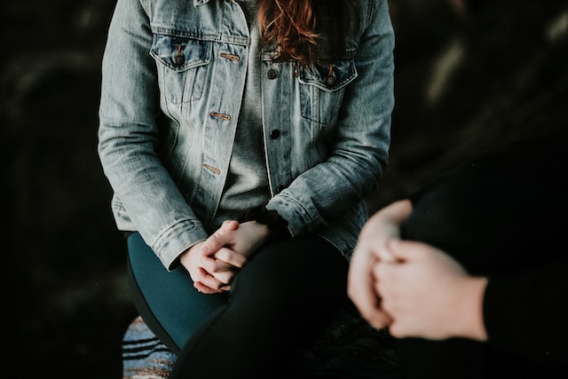 A woman in a denim jacket sits with her hands folded in a dark space. Seeking professional help with a licensed therapist can help navigate grief. Call today to get started.
