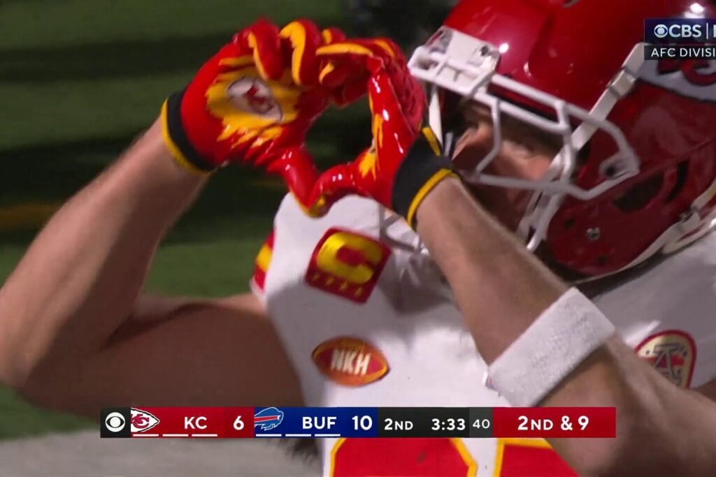 Image of Travis Kelce in football gear on the field during a game, holding his hands up in a heart shape. Get thoughts about fandom and family from a Ballwin, MO therapist here.