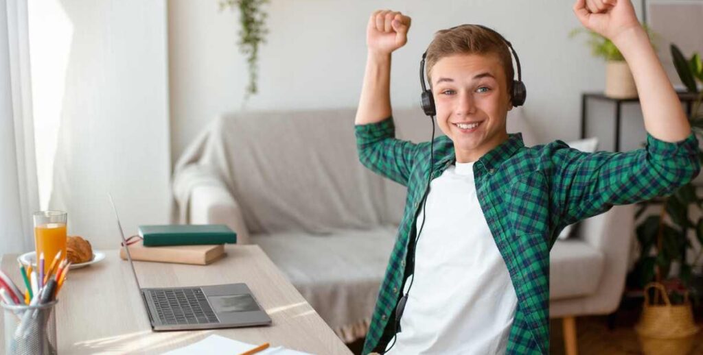 Happy teenage boy celebrating with hands raised in the air at a computer table wearing headphones. Teens can feel happy, balanced, fulfilled, and more with the help of a teen therapist near St. Louis, MO here.